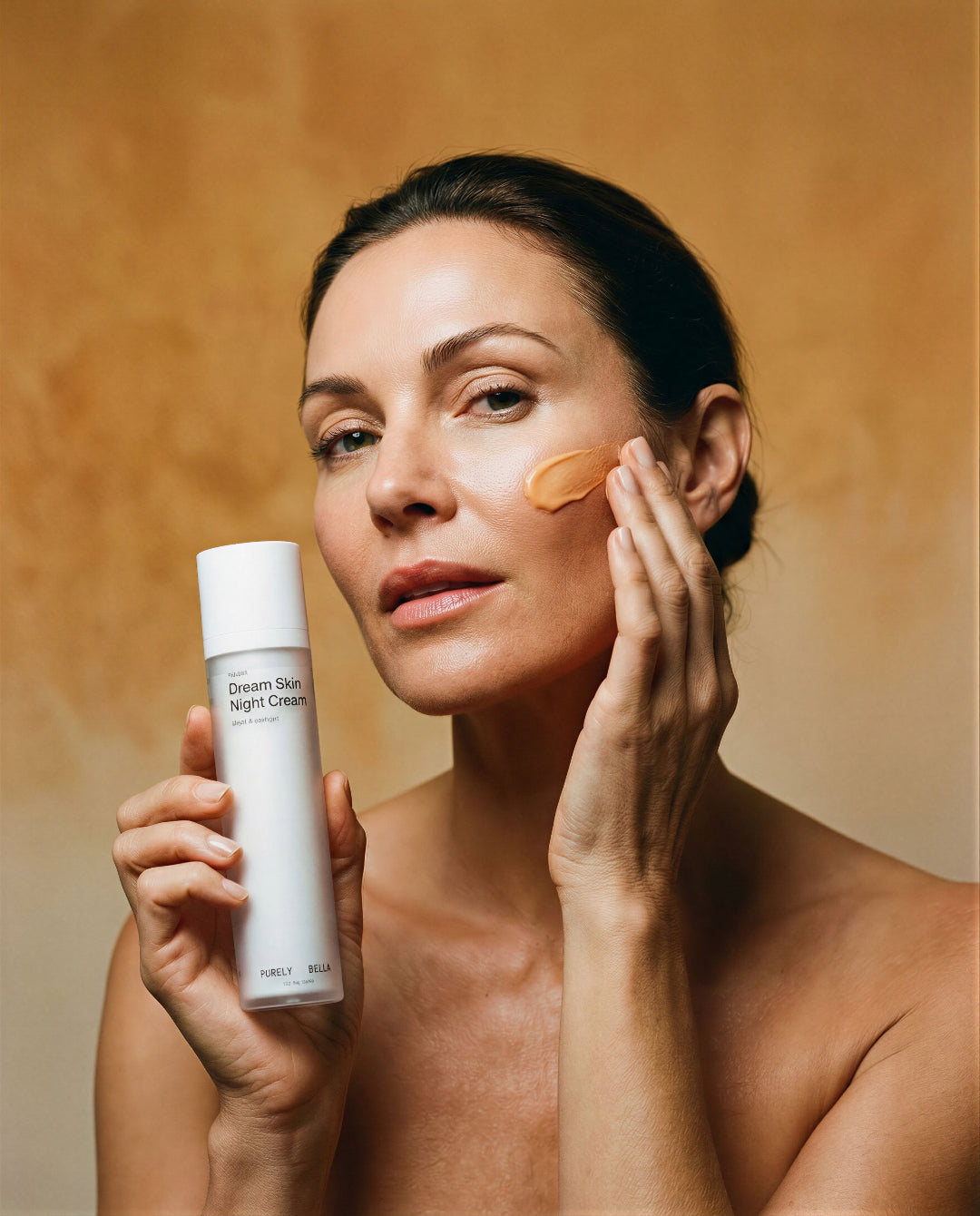Woman applying cream to her face with a skincare product in hand against a warm brown background