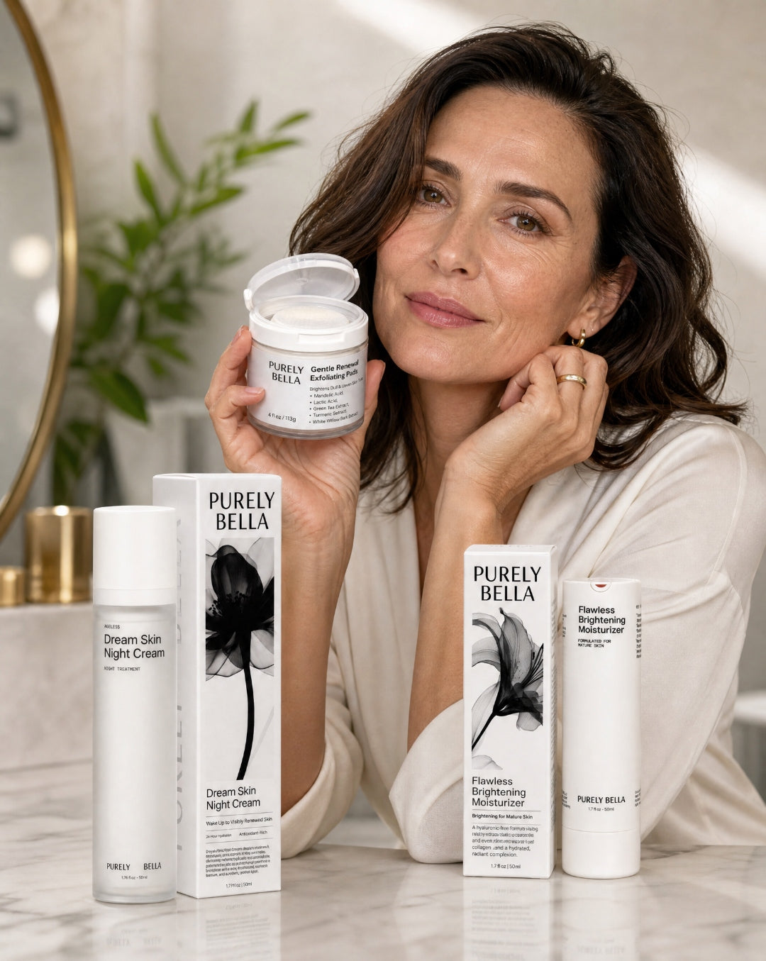 Woman in her 40s holding Purely Bella Gentle Renewal Exfoliating Pads, with Dream Skin Night Cream and Flawless Brightening Moisturizer on a marble vanity