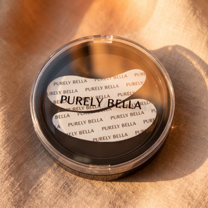 A round clear plastic compact containing black powder, with a white sticker label reading 'PURELY BELLA' repeated around the edge. The compact is photographed on a warm beige fabric surface with strong sunlight and shadows.