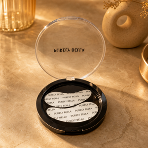An open black makeup compact with a clear lid resting on a beige stone surface. The compact interior shows white powder with repeated 'PURIFY BELLA' text, and the brand name 'PURIFY BELLA' is printed on the clear lid.