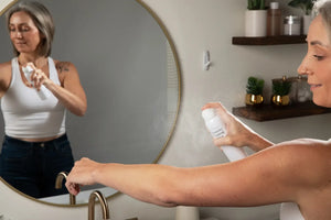 Woman applying mist to her arm in front of a mirror in a bathroom.