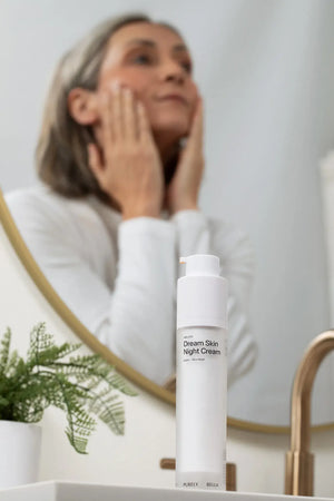 Woman applying cream to her face with a bottle of 'Dream Skin Night Cream' in front of a mirror.
